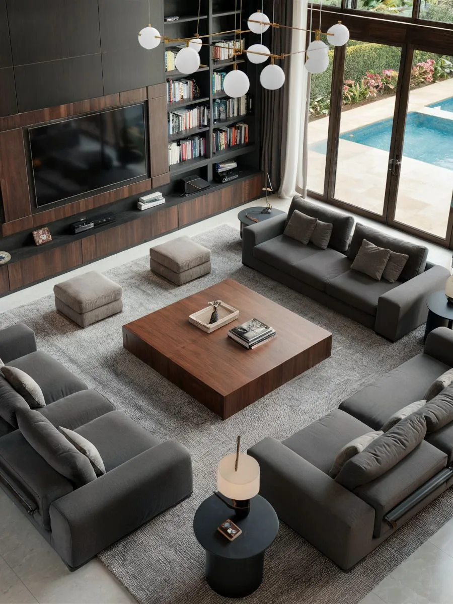 A double-height living room seen from above, a wall-mounted TV set into dark timber panelling with bookshelves, and grey sofas around a square wooden coffee table